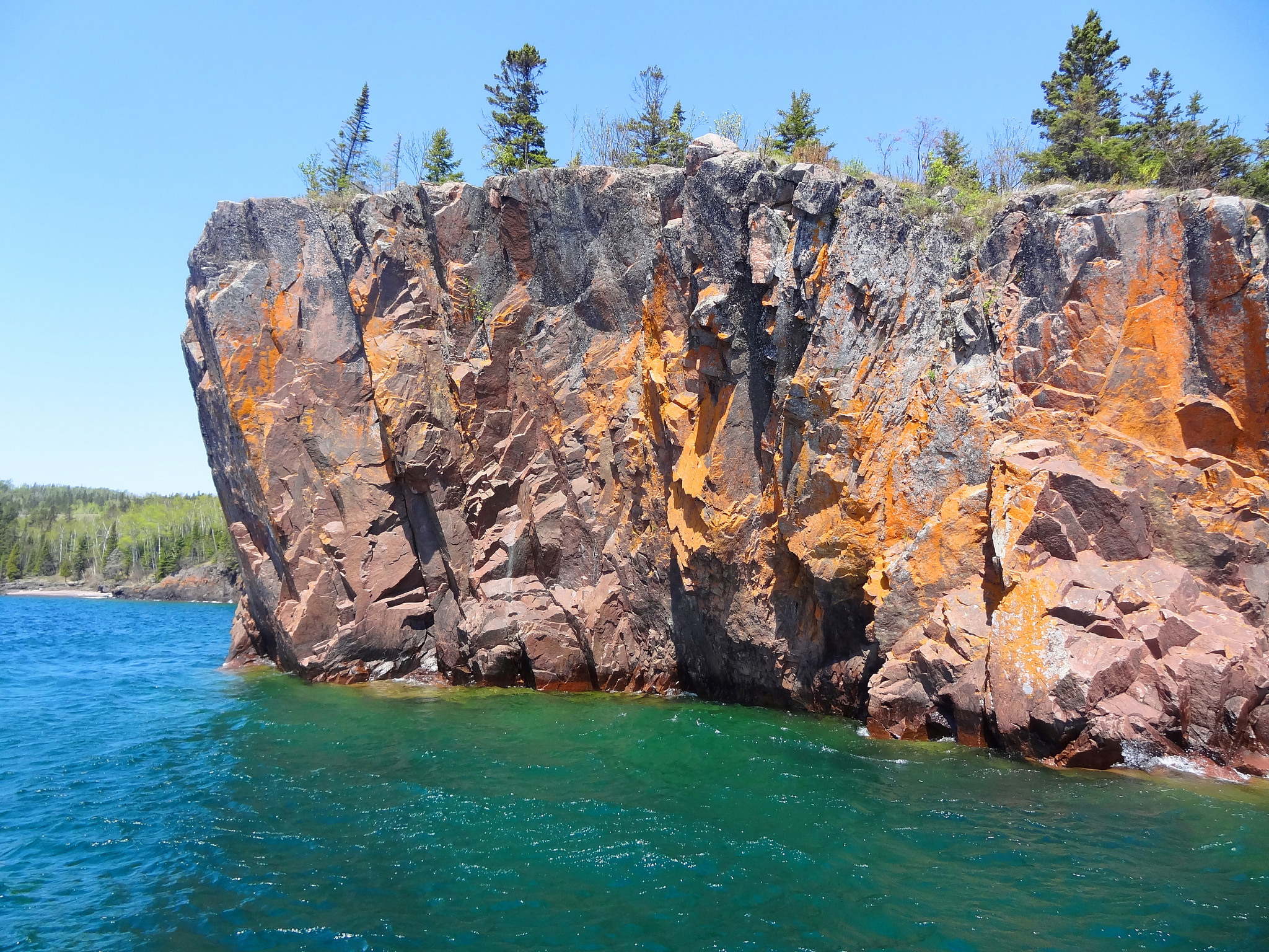 A cliff face with pine trees above blueish green water