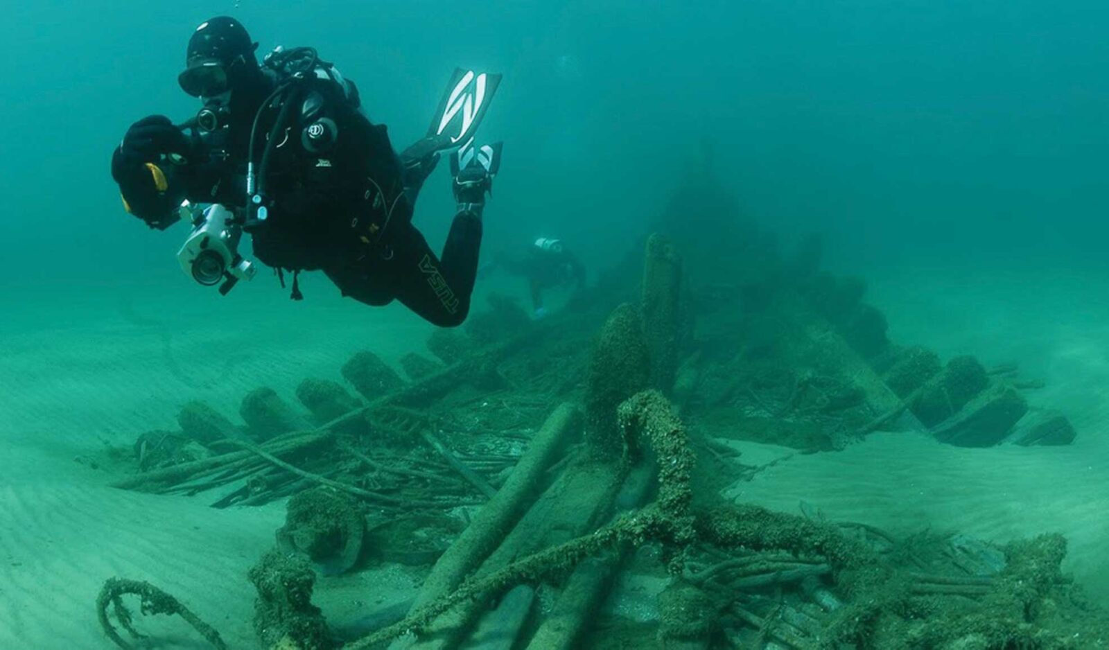 Photo of underwater maritime archaeologist swimming with a camera, near a shipwreck.